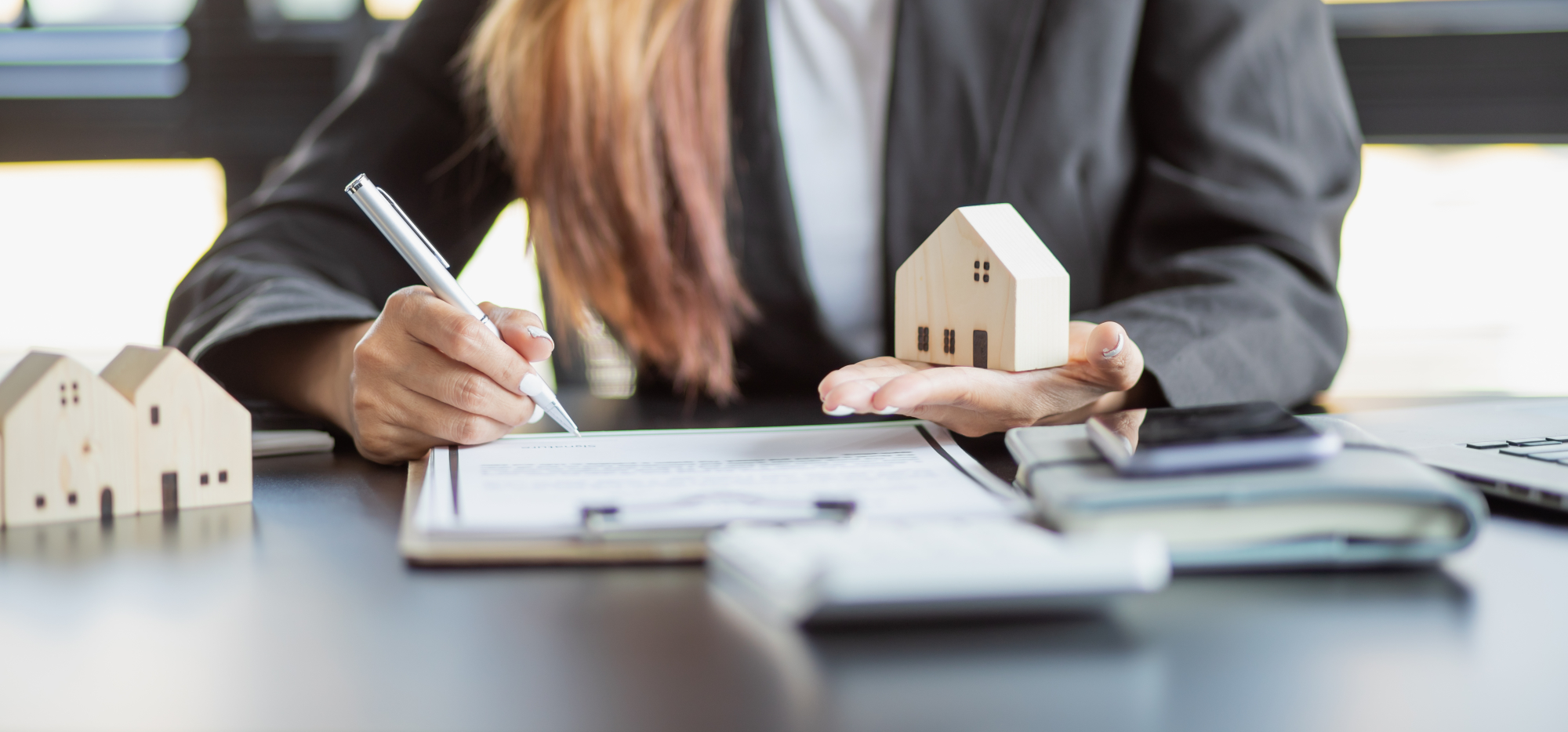 Une femme d'affaire tient une maison miniature en bois dans une main et signe un contrat avec son autre main