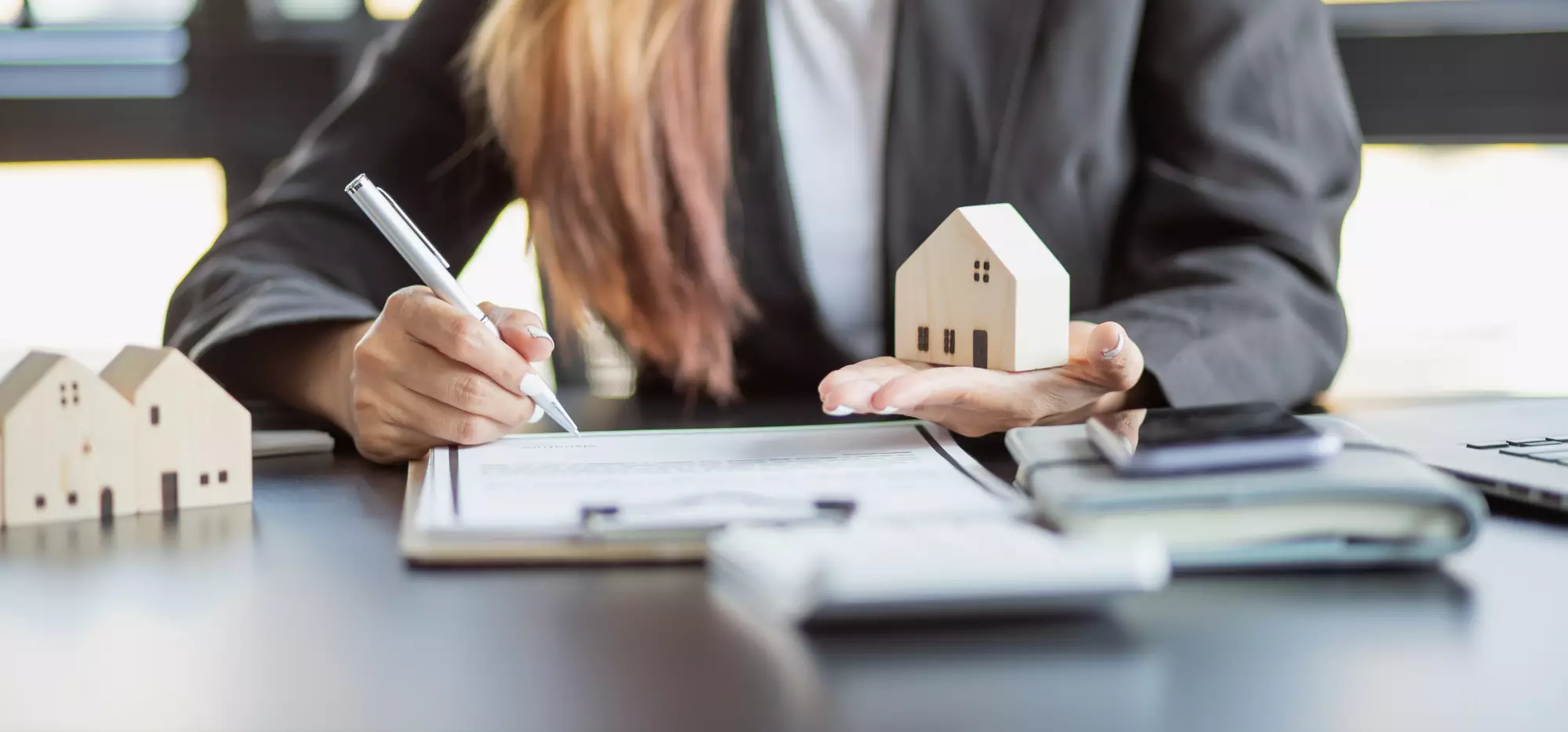 Une femme d'affaire tient une maison miniature en bois dans une main et signe un contrat avec son autre main