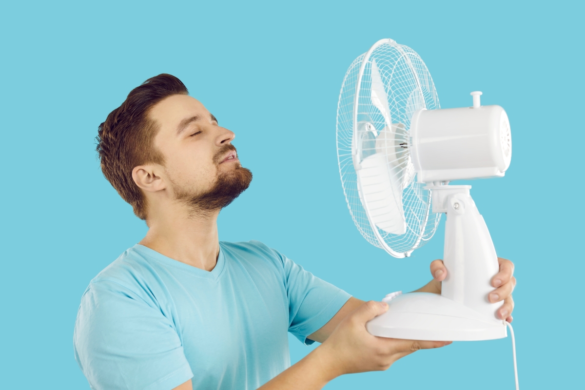 Quelques gestes simples permettent de faire diminuer de quelques degrés la température d’un logement sans avoir recours à la climatisation Bouilloire thermique — Un jeune homme tient un ventilateur devant son visage