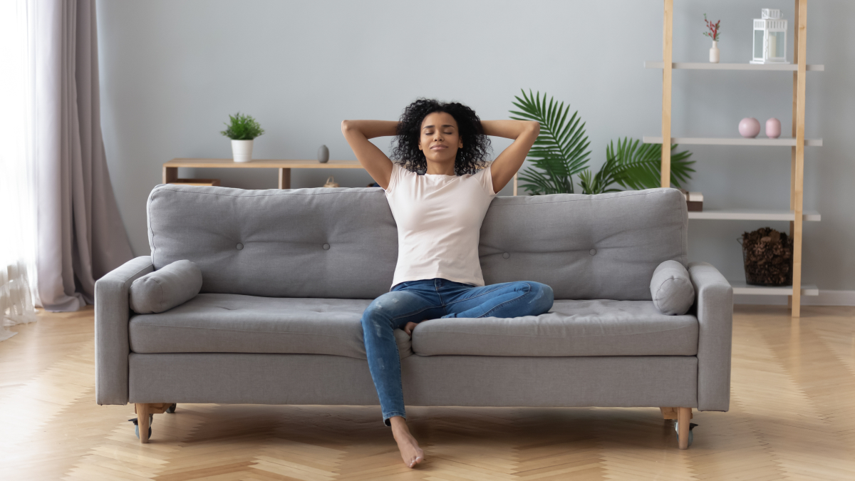 Avec la RE2020, le confort d’été est pensé dès la phase de conception des derniers programmes immobiliers neufs Bouilloire thermique — Une jeune femme à l’aise se repose sur un canapé dans un appartement neuf